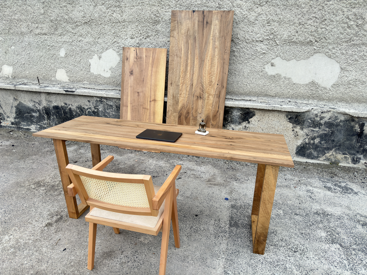 A rustic wooden writing desk with a matching wooden chair featuring a woven cane backrest, set against a textured concrete wall with additional raw wood panels.