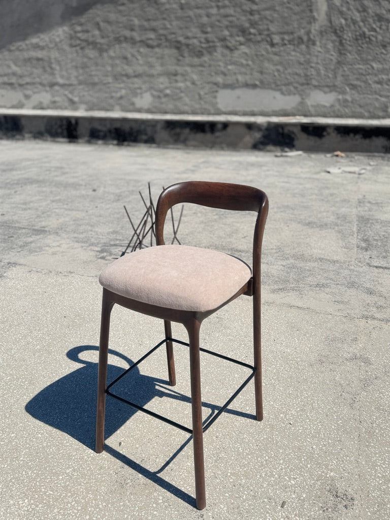 A mid-century modern bar stool with a curved dark wood frame, a light beige upholstered seat cushion, and a black metal footrest, photographed outdoors on a concrete surface.