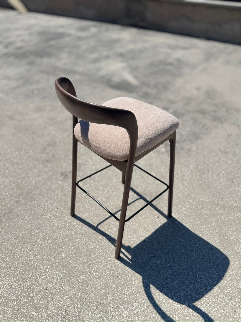 A modern bar stool with a curved dark wood backrest, a light beige upholstered seat cushion, and a thin black metal frame, photographed outdoors on a concrete surface in bright sunlight.