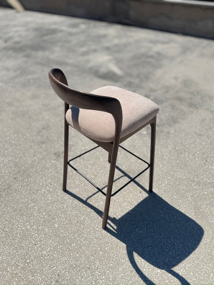 A modern bar stool with a curved dark wood backrest, a light beige upholstered seat cushion, and a thin black metal frame, photographed outdoors on a concrete surface in bright sunlight.