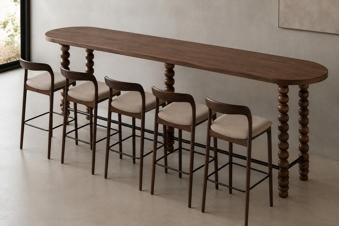 A long oval bar-height wooden table with turned spindle legs, accompanied by five matching bar stools with upholstered cream seats and dark wood frames, shown in a minimalist interior setting.