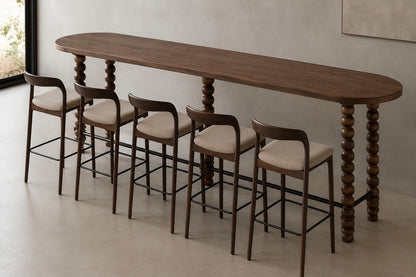 A long oval bar-height wooden table with turned spindle legs, accompanied by five matching bar stools with upholstered cream seats and dark wood frames, shown in a minimalist interior setting.