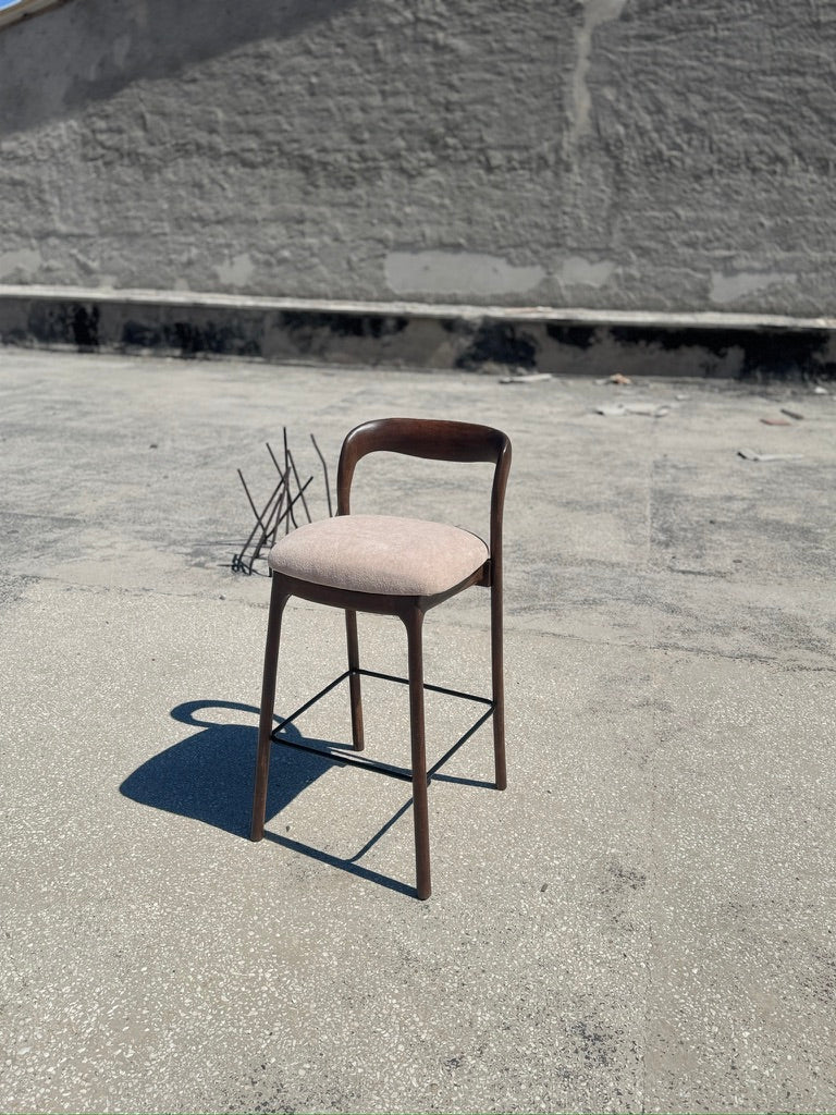 A single wooden bar stool with a curved backrest and a light beige upholstered seat cushion, photographed outdoors on a concrete surface against a textured gray wall.