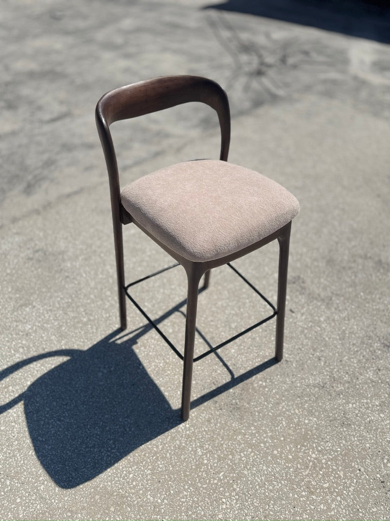 A modern bar stool with a curved dark wood backrest, a square upholstered seat cushion in a light beige fabric, and a dark metal frame, photographed outdoors on a concrete surface.